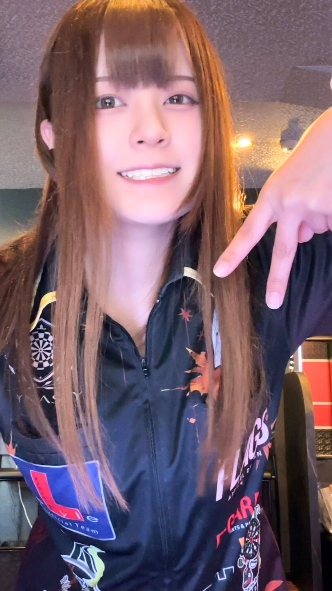 ユニフォーム姿でピースサインを見せる女性プロダーツプレイヤー中谷祐里プロ。元気いっぱいの笑顔が印象的