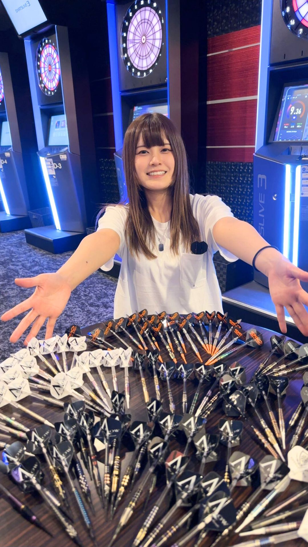 ダーツバーでたくさんのダーツを前に両手を広げて笑顔を見せる女性プロダーツプレイヤー中谷祐里プロ。明るくフレンドリーな雰囲気