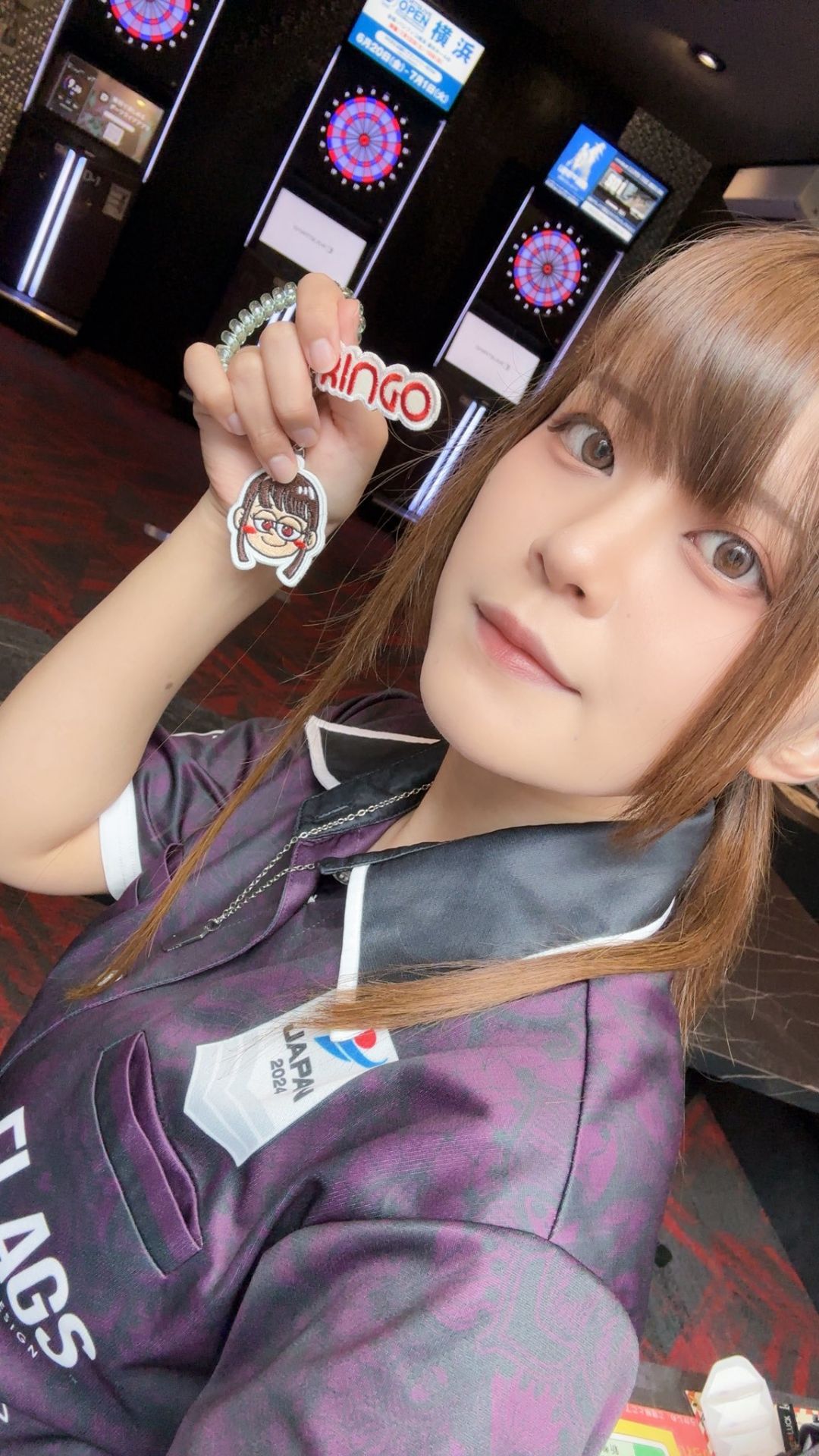 ダーツバーの前でキャラクターキーホルダーを持ち、カメラに微笑む女性プロダーツプレイヤー中谷祐里プロ