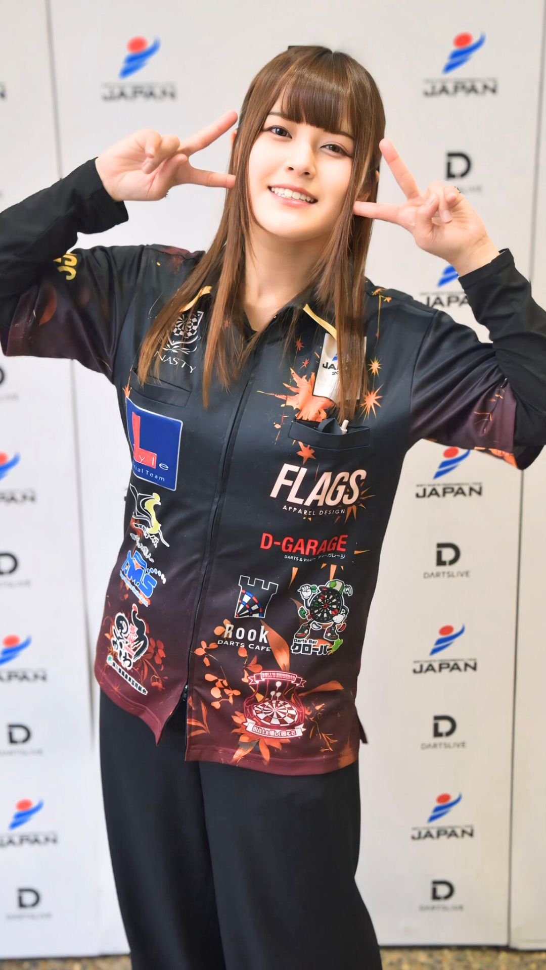 スポンサーのロゴが入ったユニフォームでダブルピースする女性プロダーツプレイヤー中谷祐里プロ。大会会場での記念撮影