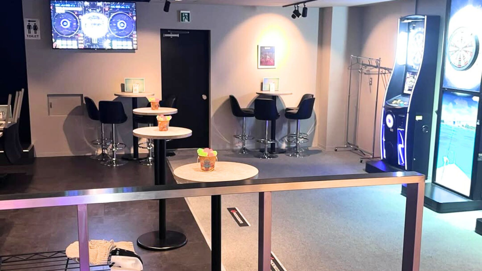 モダンなバーの店内。複数のダーツマシンが並び、高いテーブルと椅子が配置されている。壁にはトイレのサインや緊急出口のサインが見える。