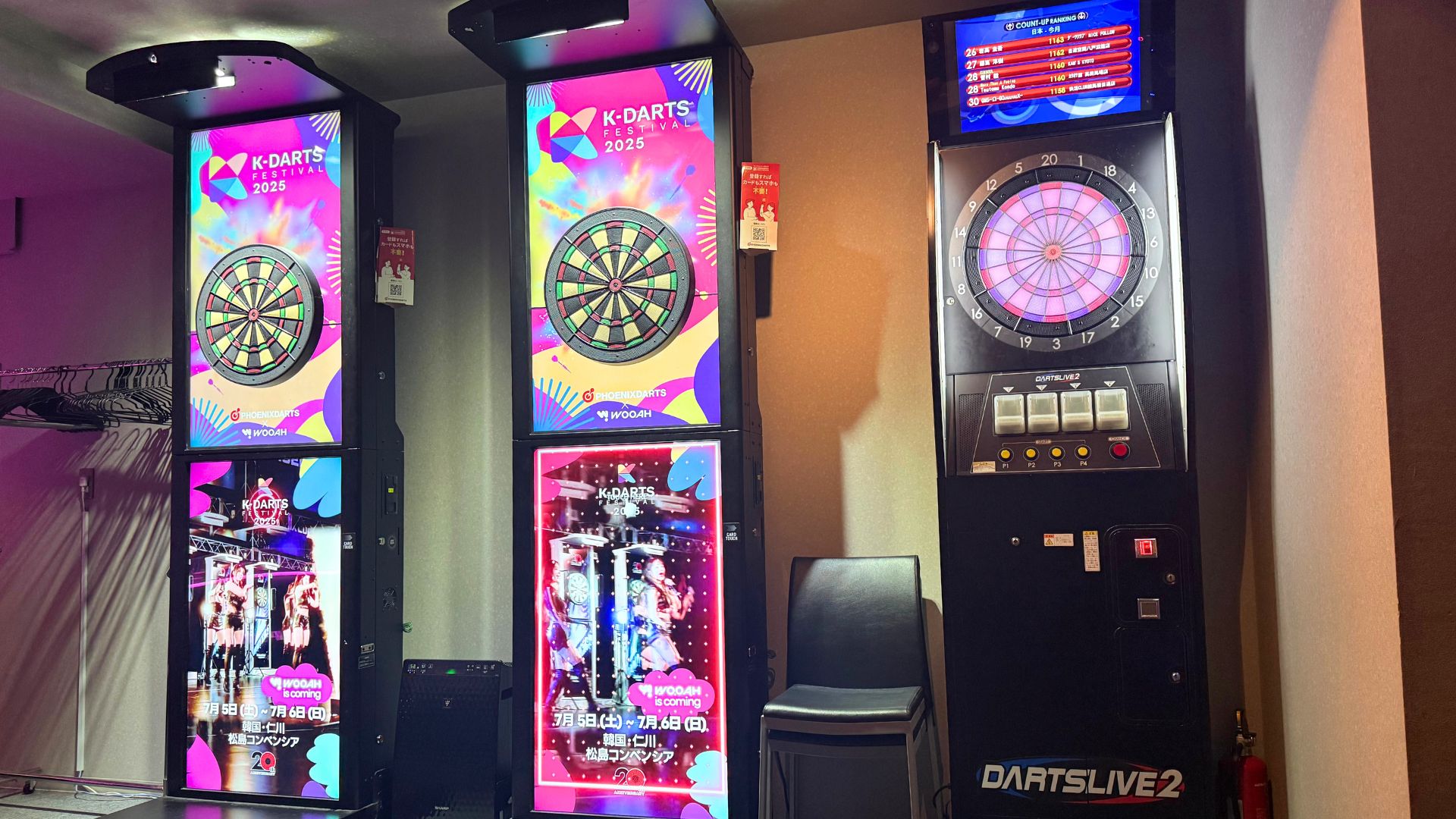 ダーツマシンが2台並んでいる様子。画面にはカラフルなダーツボードとイベントのポスターが表示されている。