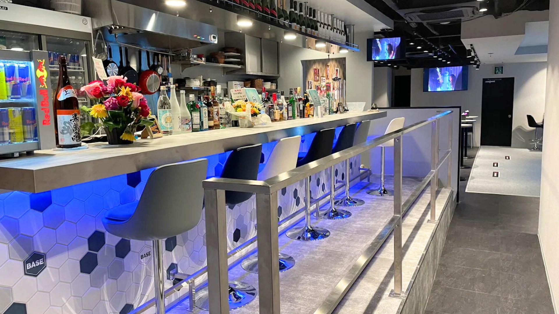 バーカウンターの内部。背後にはボトルが並び、カウンターは青い光で照らされている。手前にはハイスツールが配置され、ドリンクが置かれている。