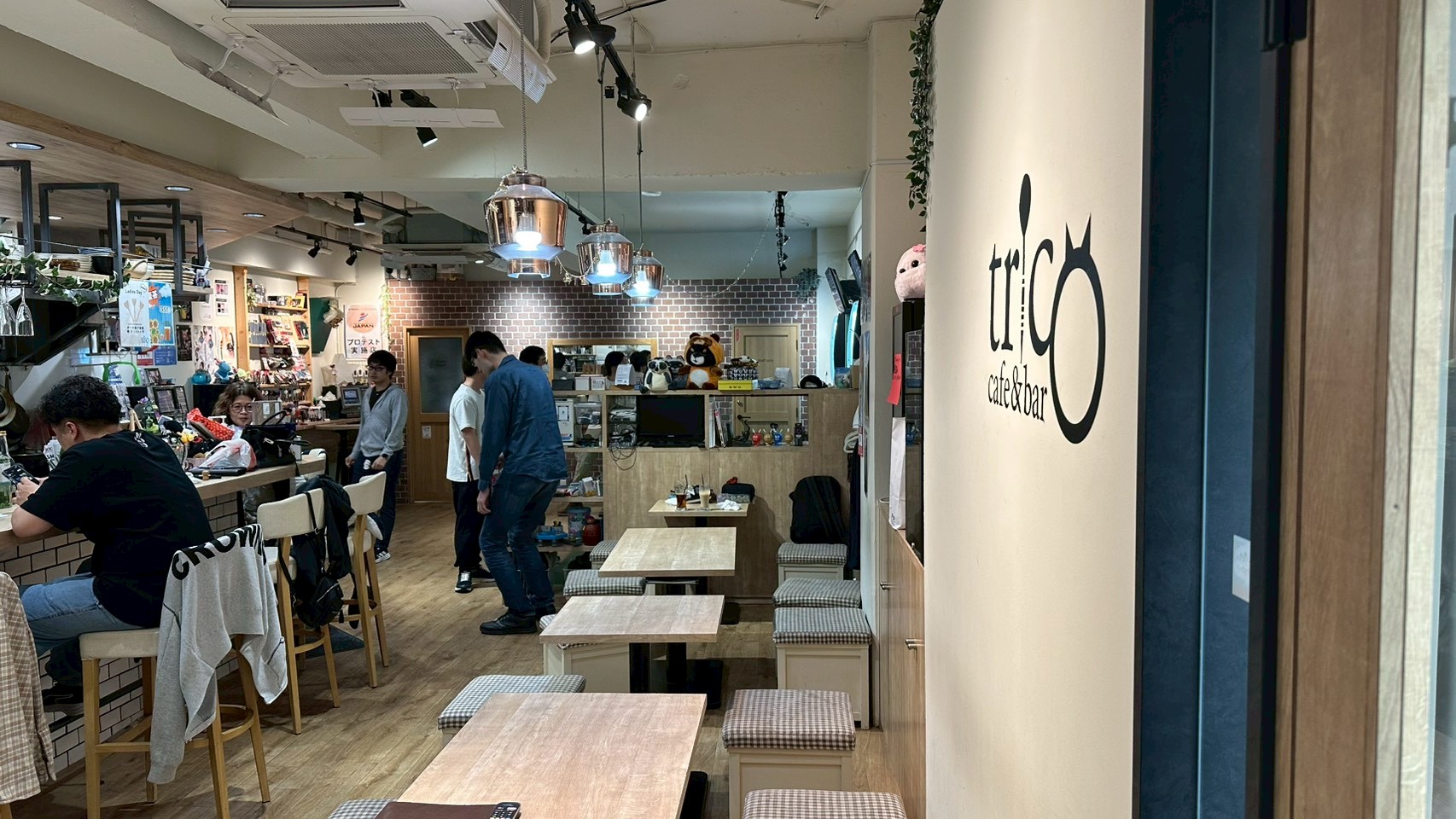 Cafe&bar tricoのおしゃれな店内風景。壁にはお店のロゴがあり、カウンター席やテーブル席でリラックスするお客さんの様子。